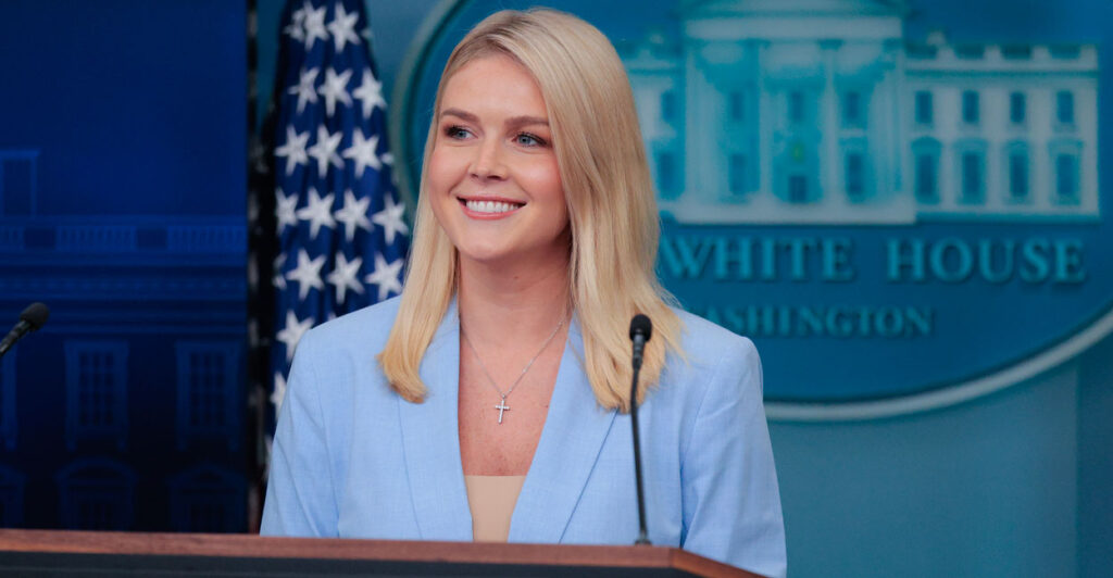 Karoline Leavitt in a robin blue jacket smiling behind podium in White House Press Briefing Room.