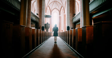 A woman in a dark, empty church walks toward an altar lit with sunlight.