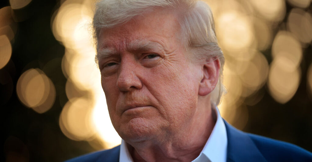 Close-up of Donald Trump in a blue jacket with a white open collar against a backdrop of diffused lights.