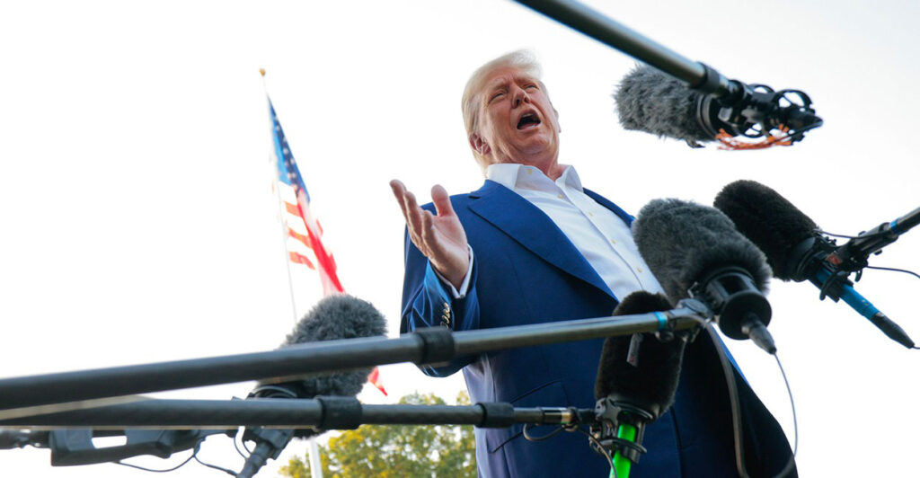 Donald Trump stands and holds his right hand out while he is surrounded by microphones.