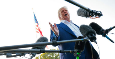 Donald Trump stands and holds his right hand out while he is surrounded by microphones.