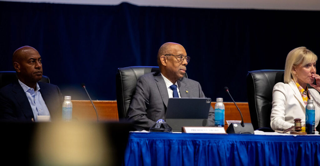 Michael Drake sits between two other University of California regents at a board meeting as he looks to his left.