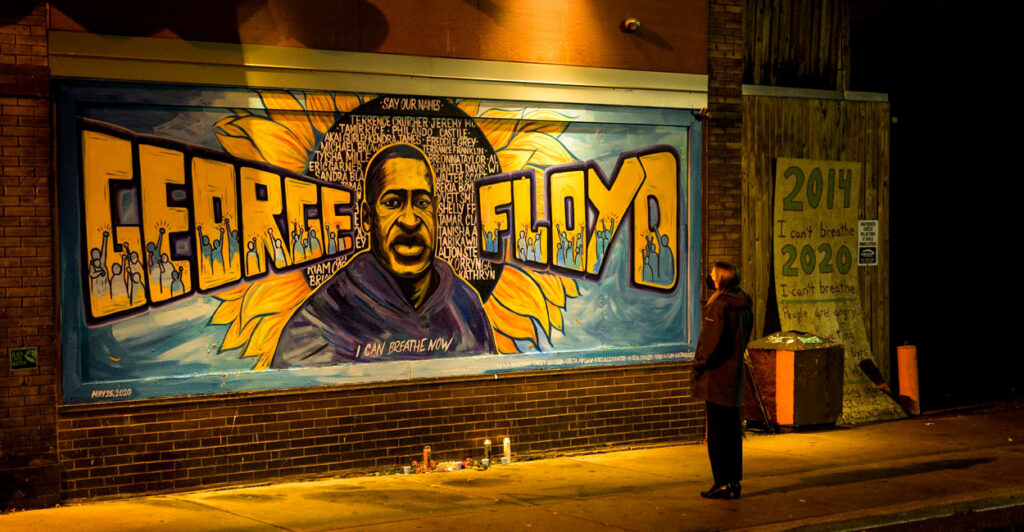 A man stands in front of a mural of George Floyd.