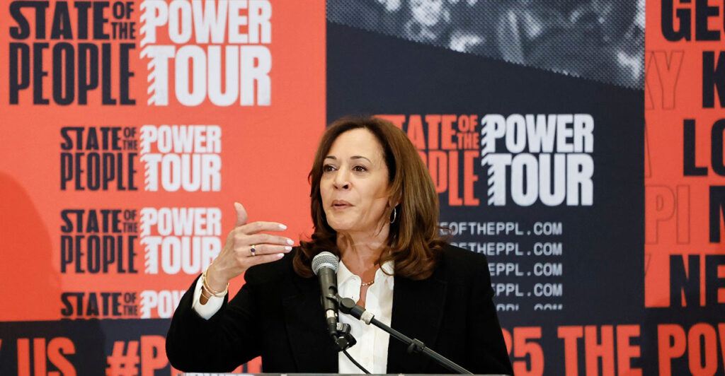 Kamala Harris stands at a podium in front of a microphone and talks while gesturing with her right hand.