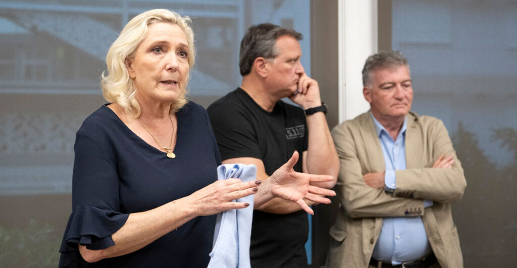Marine Le Pen stands gesturing with her hands while talking to a crowd of reporters.