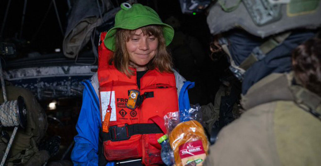 Greta Thunberg in an orange life-preserver and green hat is handed a sandwich.