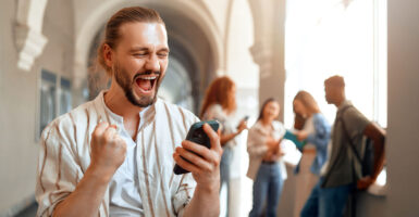 An excited male college student with a beard reacts to good news on smartphone.
