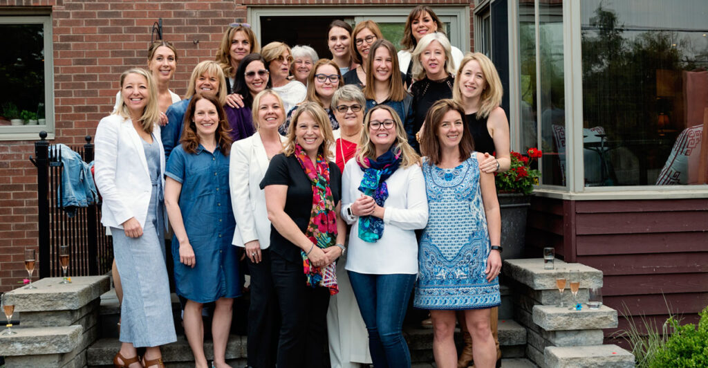A group of happy women aged 18-70 stand together on the steps in front of a brick building. On the side of the steps glasses of champagne.