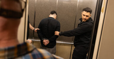 Two plainclothes ICE agents place an arrested immigrant into an elevator. Suspect's back is to the camera.