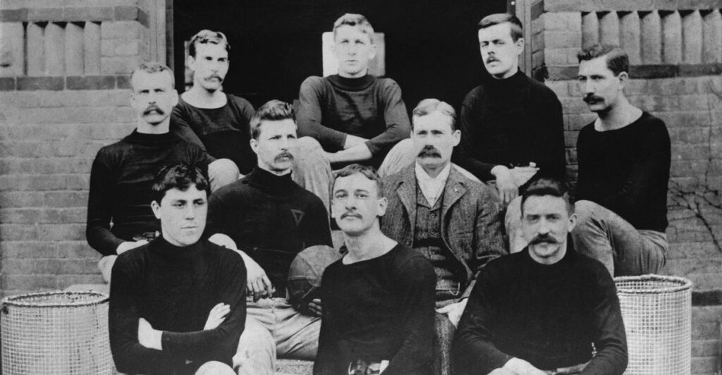A 1891 shot of James Naismith in a suit sitting on steps with the first basketball team. The young men decked in black long sleeve shirts and tan trousers.