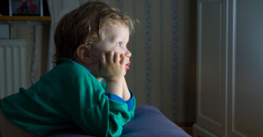 Adorable little boy in green long sleeve shirt, head in hands, stares in rapt attention at an unseen TV.