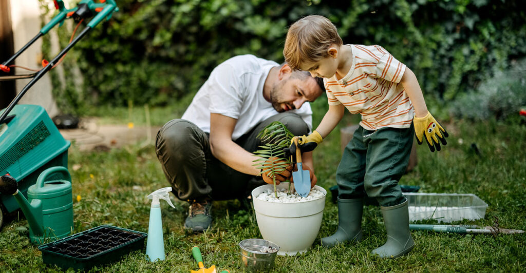 A father and son work together to plant a small tree in a white flowerpot.