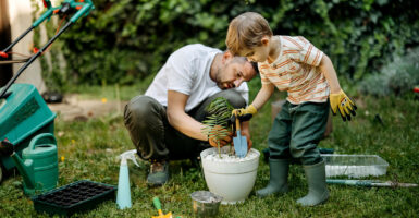 A father and son work together to plant a small tree in a white flowerpot.