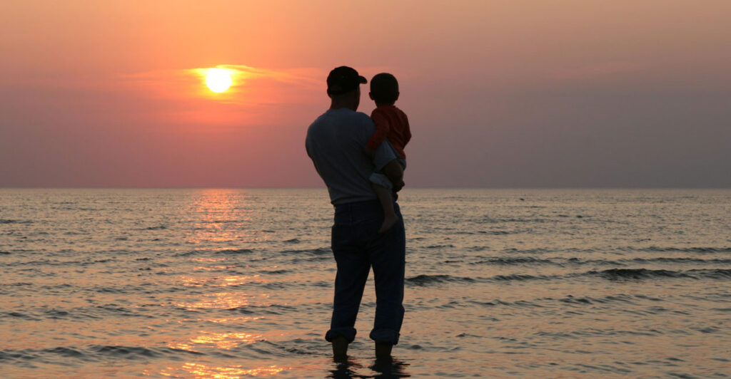 A silhouette of a father holding his son at the shoreline watching a sunset.