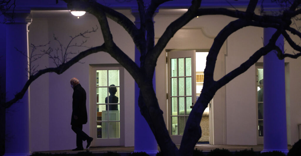 President Joe Biden in silhouette leaves Oval Office at night, as lights remain on and woman stands, presumably still working.