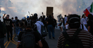 Shot from behind, a swarm of anti-ICE, anti-Trump protesters moves toward police in a street filled with smoke.