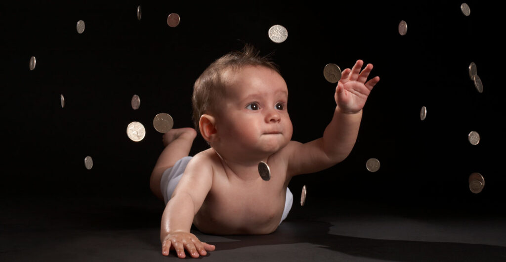Infant boy on his belly against a black background reaches in the air as coins fall around him.