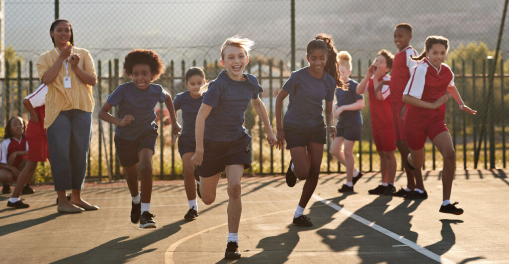 A group of young girls in gym clothes run toward the camera, as several classmates look on.