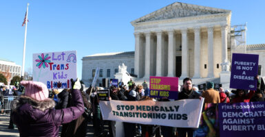 Demonstrators in support of a ban on "gender-affirming care" for minors stand in front of Supreme Court with signs and banners. One pro-gender-affirming care demonstrator faces them with her own sign reading "Let Trans Kids Bloom."