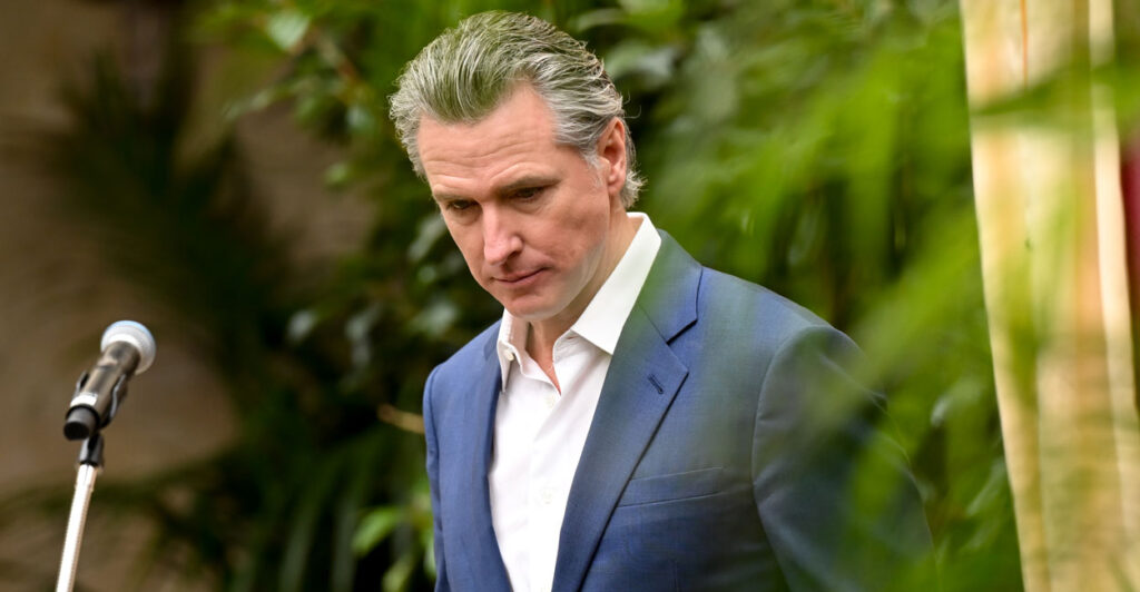 Gavin Newsome, in a blue jacket and open white shirt looks down during an outside event.