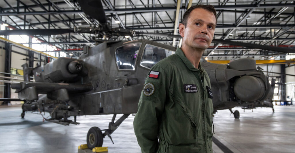 Polish Major Mariusz Strzyzewski from The 1st Aviation Brigade stands in uniform with his hands behind his back in front of an AH-64D APACHE helicopter.