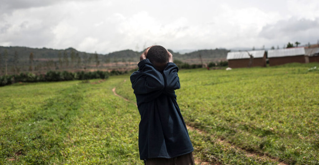 A Nigerian man in a field holds the back of his head with both hands, arms covering his face in grief.