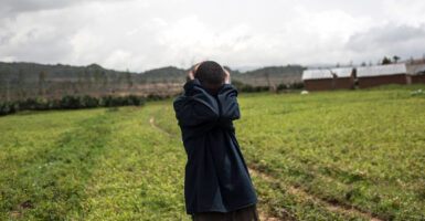 A Nigerian man in a field holds the back of his head with both hands, arms covering his face in grief.
