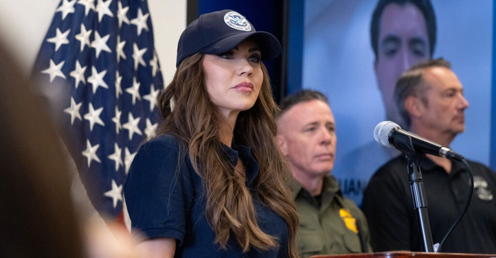 Kristi Noem in a blue short-sleeve shirt and blue hat listens at a press conference., with two men behind her.