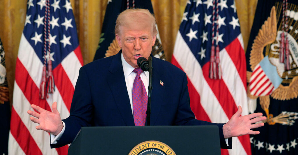 Donald Trump, arms spread behind a podium, with flags behind him.