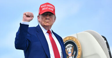 President Trump raises his fist while boarding Air Force One and waring a MAGA hat, red tie, and dark blue suit.