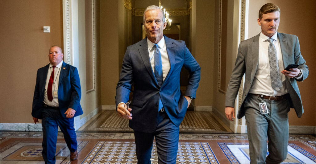 Senate Majority Leader John Thune strolls down a Senate corridor, flanked by two other people.