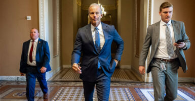 Senate Majority Leader John Thune strolls down a Senate corridor, flanked by two other people.