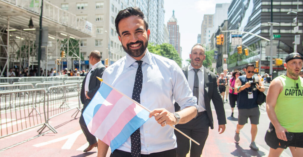 Zohran Mamdani at a NYC Pride parade holding a blue, white and pink transgender flag.