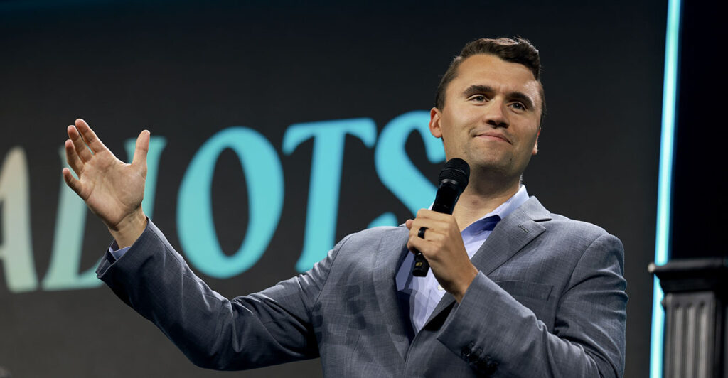 Turning Point USA Founder Charlie Kirk in a grey suit gestures with a microphone