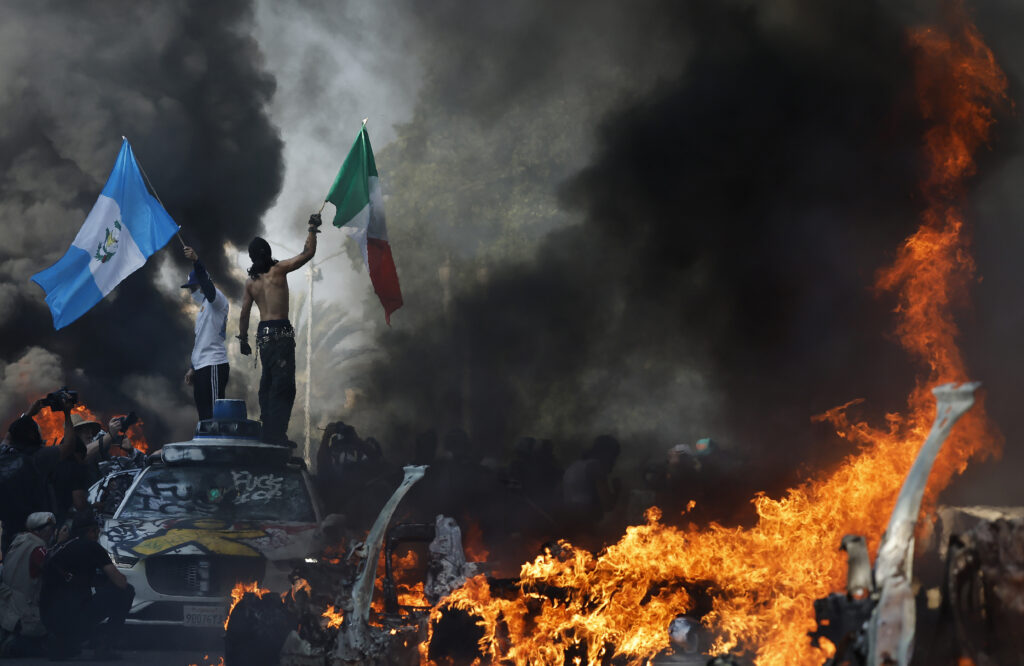 Two men wave the Mexican and Guatemalan flags amid burning cars during the LA riots.