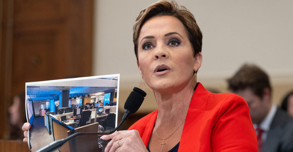 Kari Lake in a red suit holding up a photo testifying before a congressional committee