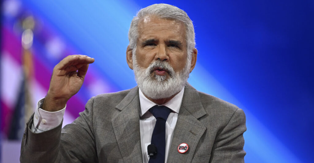 Dr. Robert Malone in a gray suit and a blue tie wears a "No World Health Organization" pin as he gestures while speaking on stage