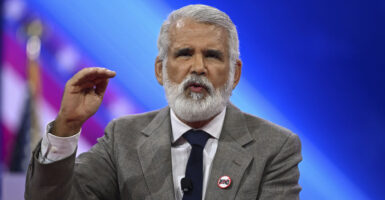 Dr. Robert Malone in a gray suit and a blue tie wears a "No World Health Organization" pin as he gestures while speaking on stage