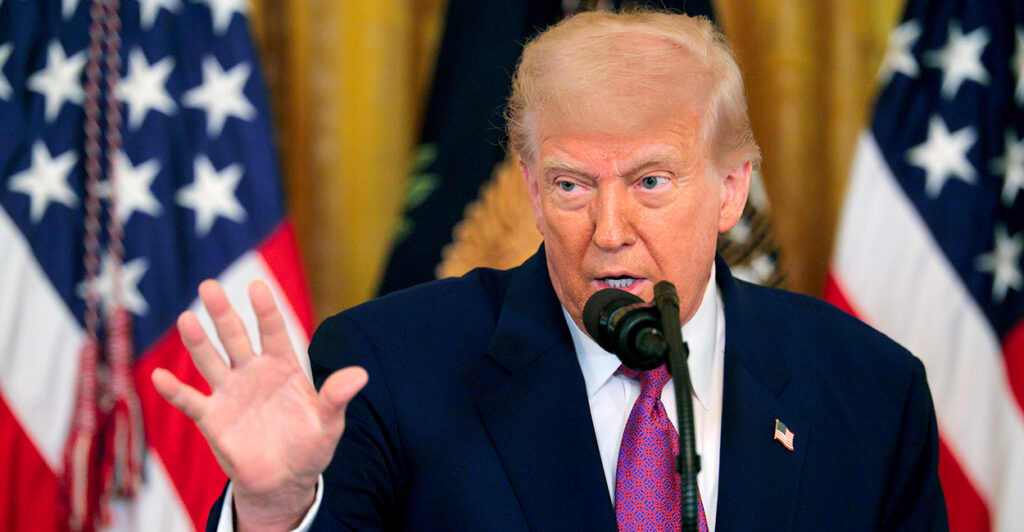 President Trump holds his hand up while speaking in front of two American flags.