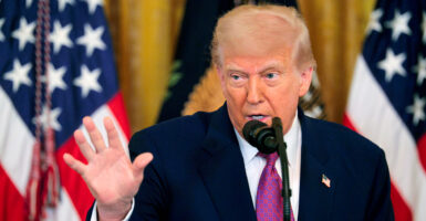 President Trump holds his hand up while speaking in front of two American flags.