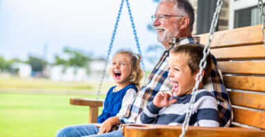 a grandfather in a plaid shirt sits laughing with his two young grandchildren on a porch swing