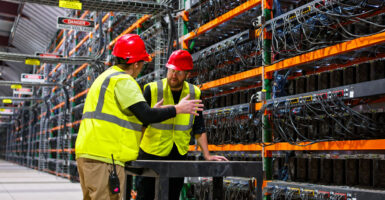 two employees in yellow fluorescent vests and red hardhats have a discussion inside a data center with thousands of computer servers, wires, and "danger -high voltage" signs