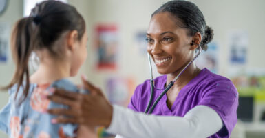 a black nurse in purple scrubs listens to the heartbeat of a young girl with a stethoscope