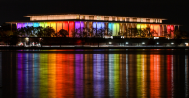 The exterior of the Kennedy Center illuminated in rainbow colors at night, reflecting on the water below it.