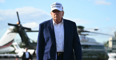 President Donald Trump in blue jacket, white shirt and white MAGA hat walks toward camera, with Marine One behind him.