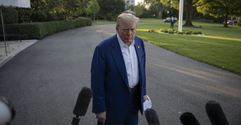 President Donald Trump faces reporters in the White House driveway, wearing a blue jacket and open collar white shirt.