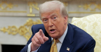 Donald Trump sits in a chair and points his right index finger at reporters.