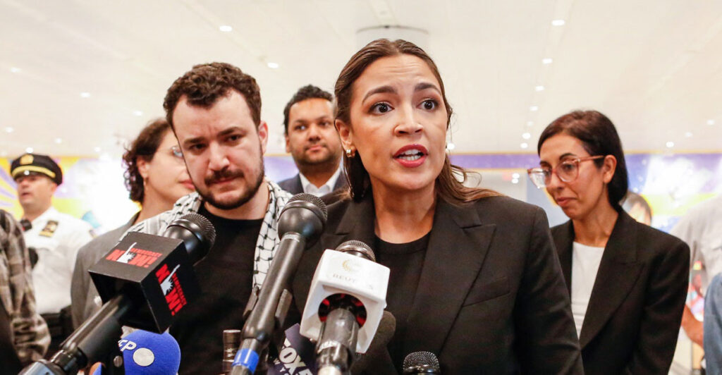 Alexandria Ocasio-Cortez stands at a podium in front of a microphone and looks to her left as she speaks.