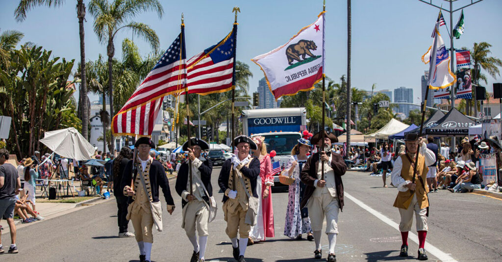 A group of men dressed like Minute Men from the Revolutionary War march down the street in a parade holding the American flag.