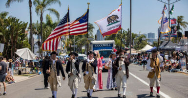 A group of men dressed like Minute Men from the Revolutionary War march down the street in a parade holding the American flag.